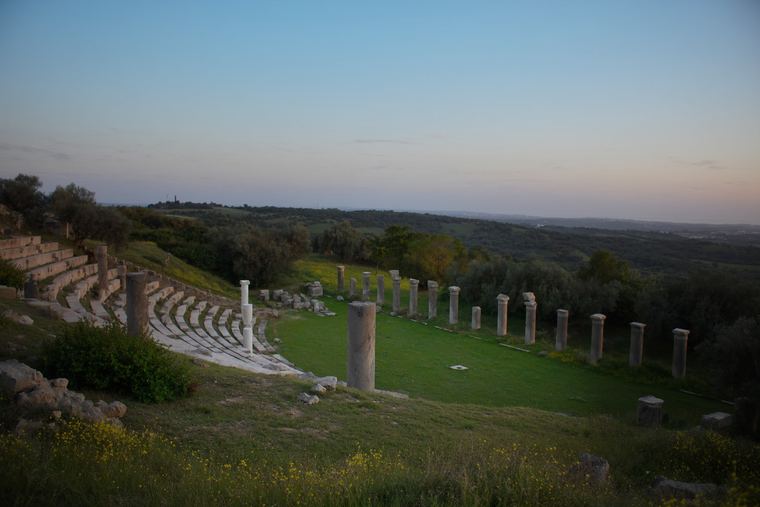 The stone seats of an ancient theatre and a row of standing columns on a grassy hillside at dusk