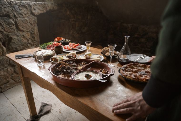 A dimly lit table in a stone house set with a baked lamb dish, a filo pie, raki glasses and wine