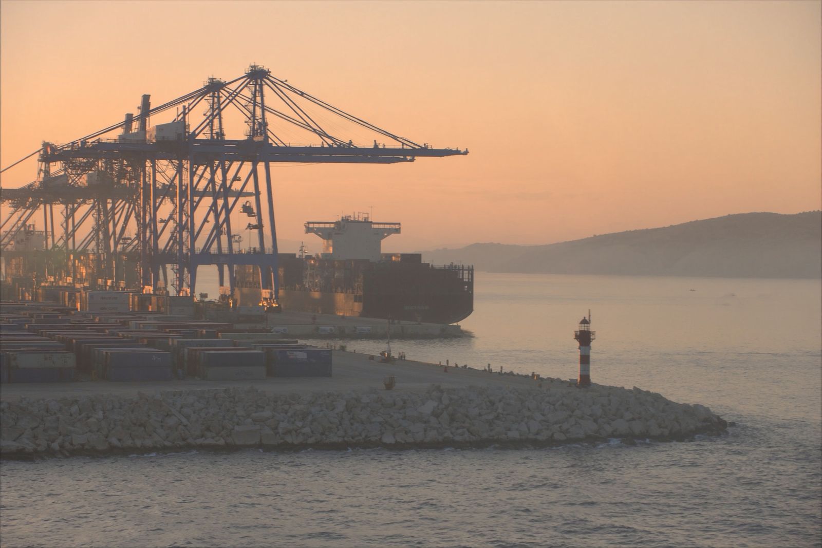 Container cranes, stacked containers and a cargo ship at a breakwater port at sunrise
