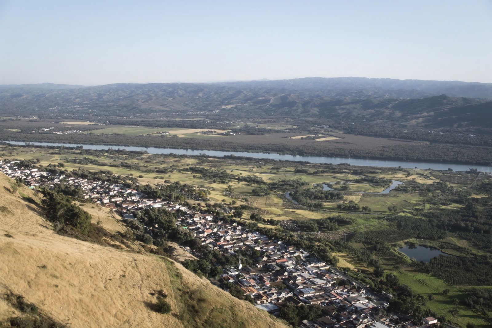 A wide river plain of fields and orchards seen from a hillside, with a town at the foot of the hills