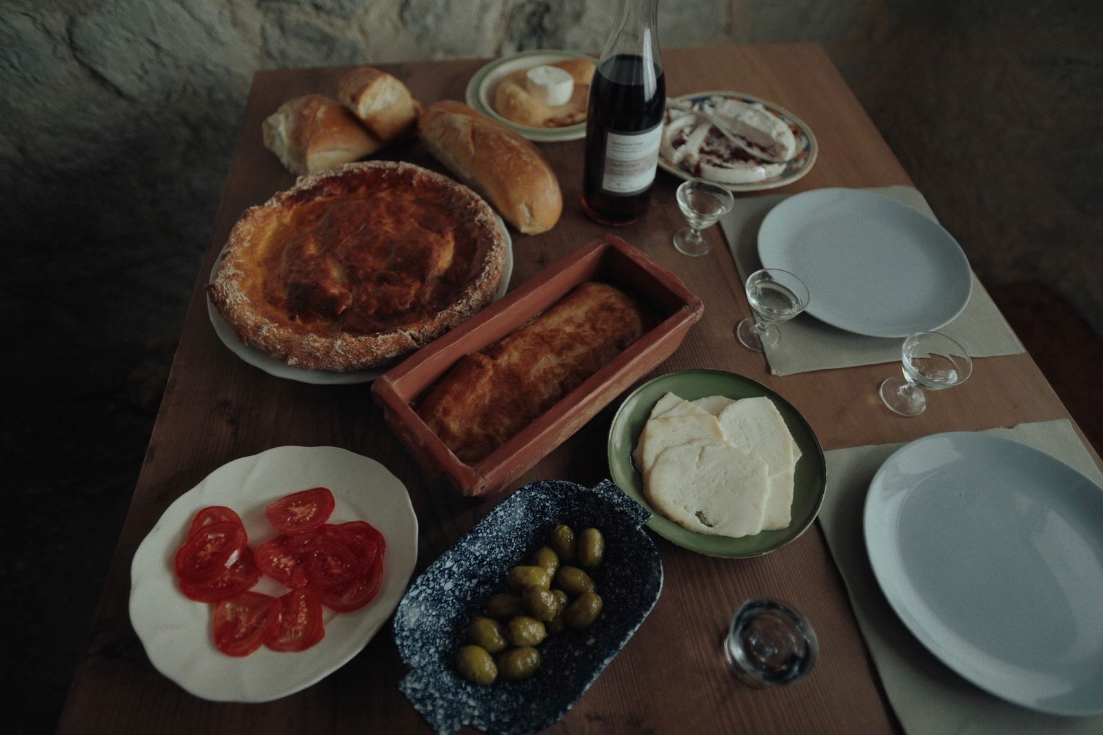 A table of home cooking seen from above: a round filo pie, a baked dish, white cheese, tomatoes, olives, bread and wine