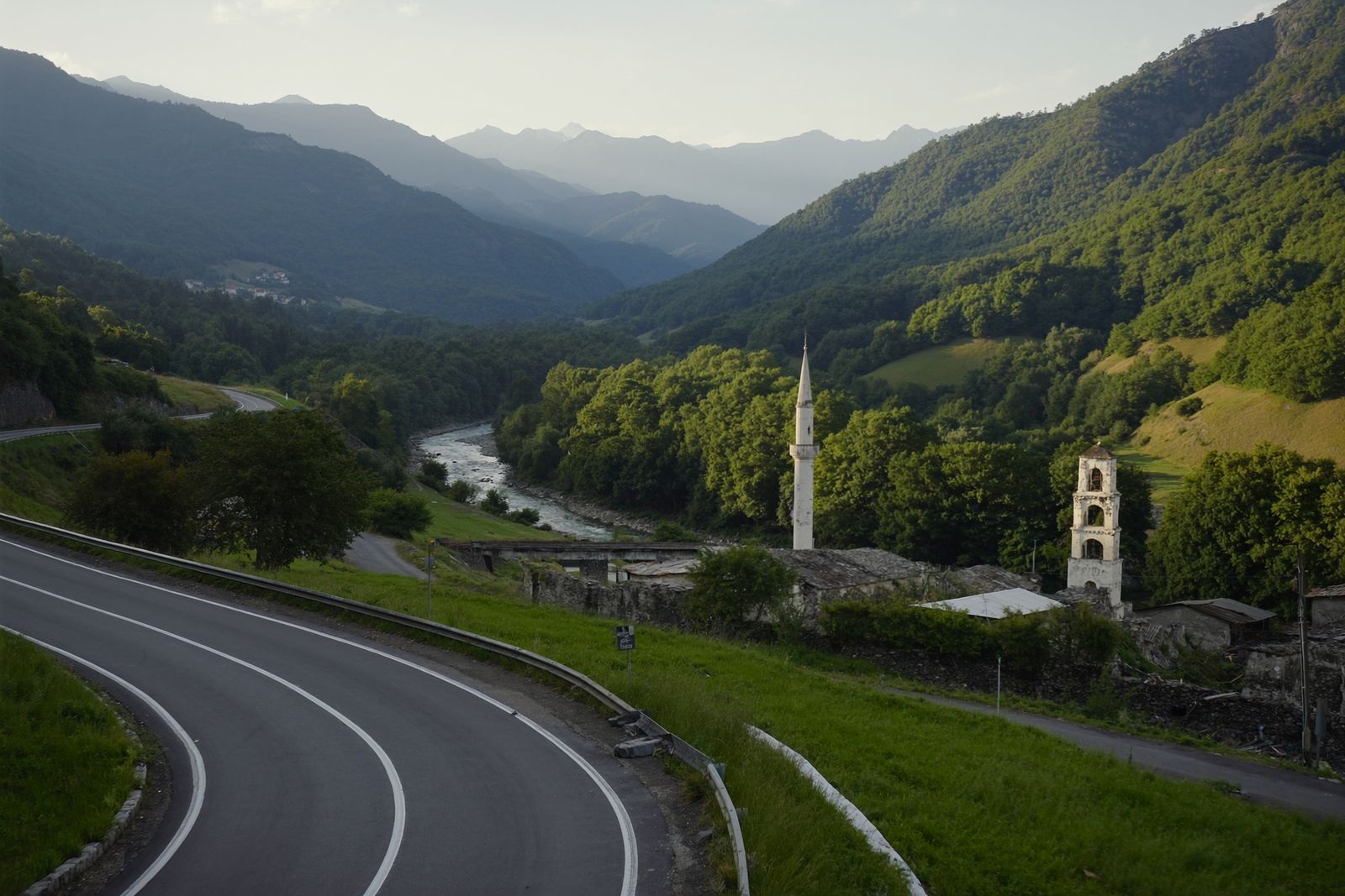 A winding road above a green river valley, with a village minaret and a church bell tower among the trees