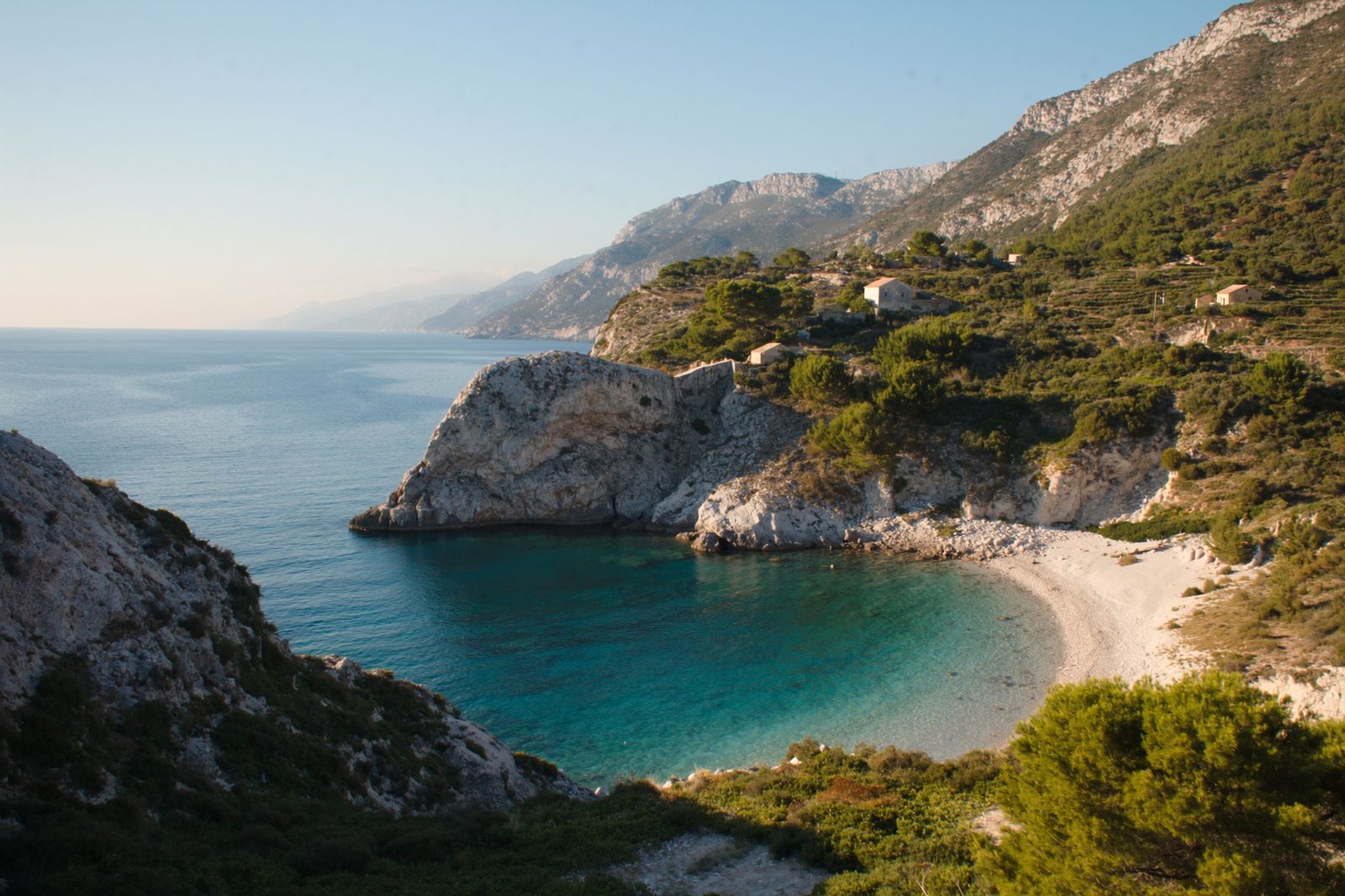 Limestone cliffs and olive terraces above a small pebble cove of clear turquoise water, with a mountain ridge running down to the sea