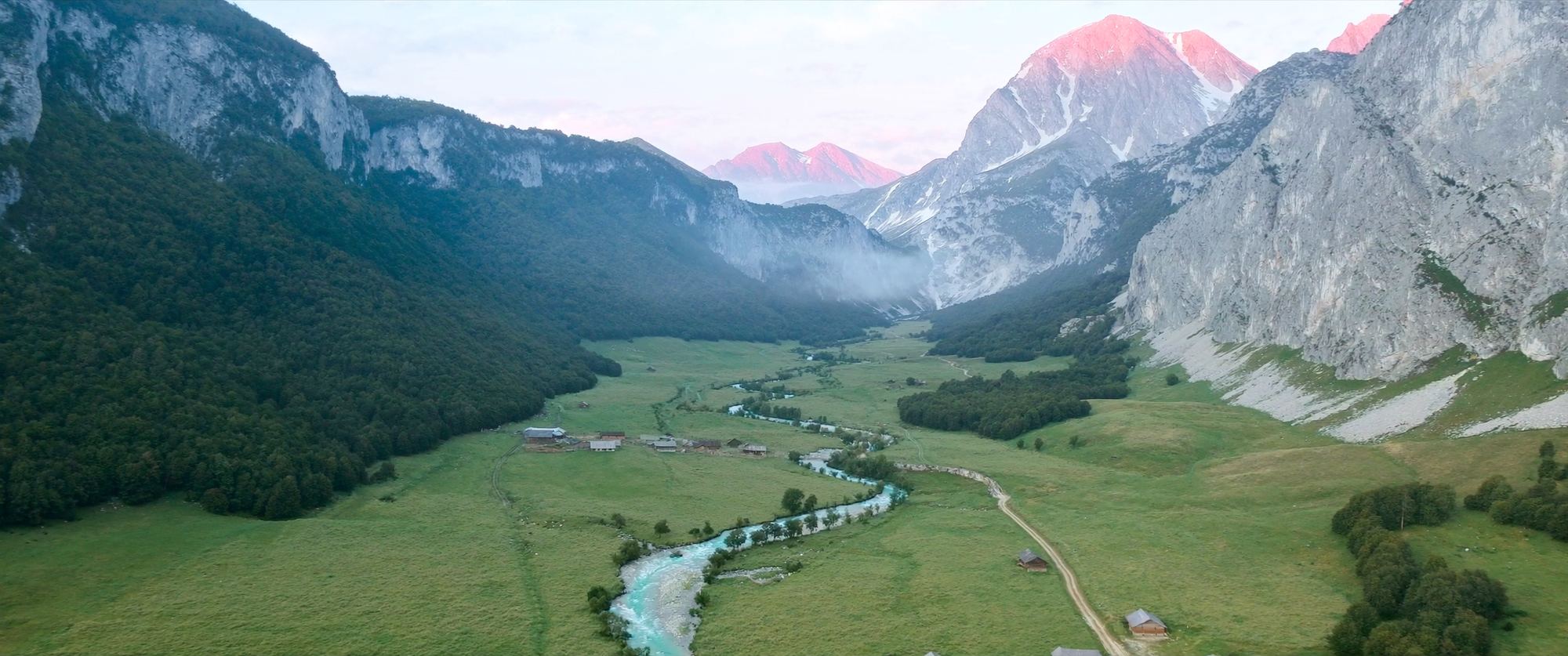 A green glacial valley with a turquoise river winding past hay meadows, beneath pale limestone peaks lit pink at dawn