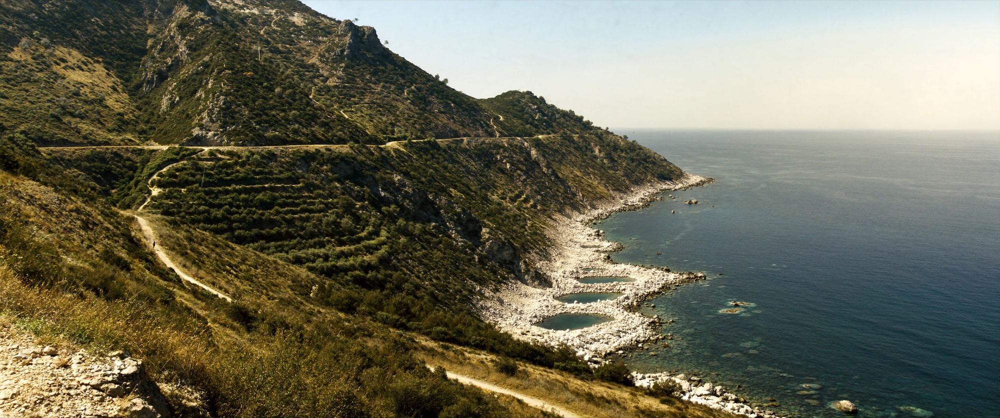 Terraced hillsides and a winding track above white pebble coves and a calm blue sea