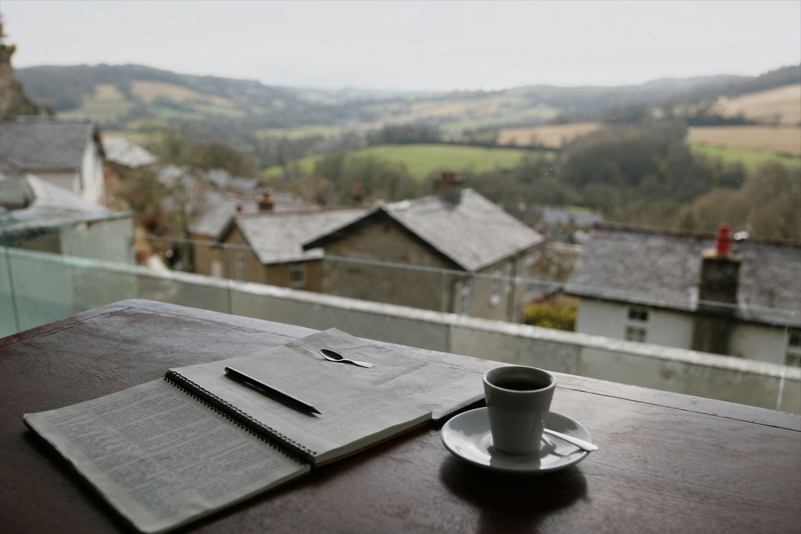 An open notebook, a pencil and a small cup of coffee on a cafe table by a window over stone-roofed houses