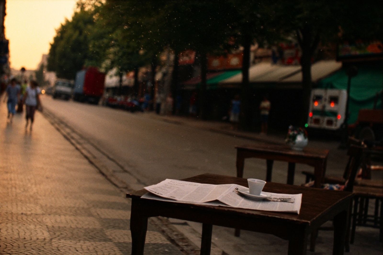 A folded newspaper and an espresso on a cafe table on a tree-lined street in the evening