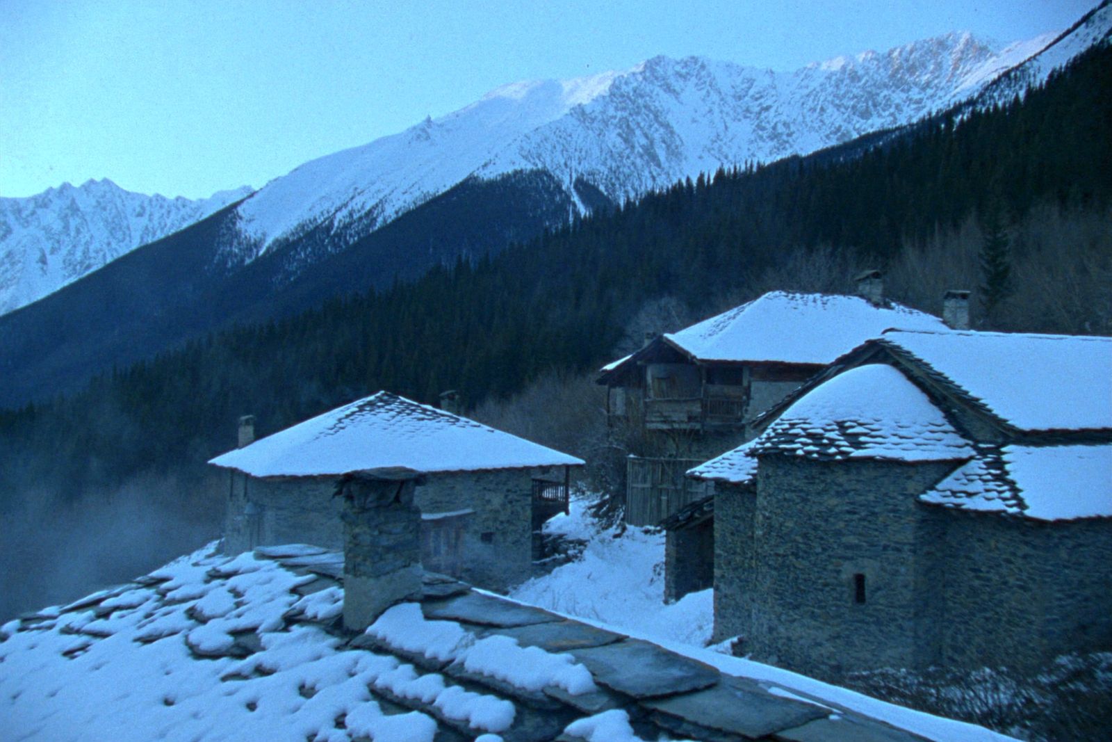 Stone houses with stone roofs under fresh snow, with pine forest and snowy peaks behind
