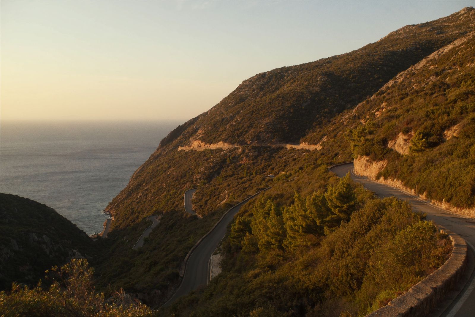 A narrow coastal road with hairpin bends cut into a mountainside high above the sea at sunset